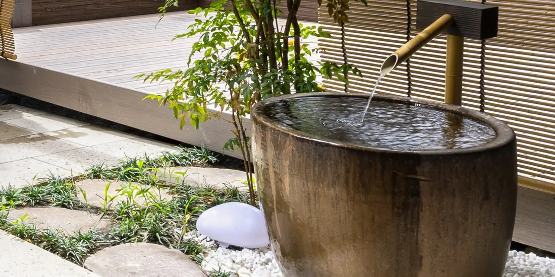 森小屋の坪庭で、竹の筧から青銅の水盤へ落ちる水。傍らには木のデッキ。
