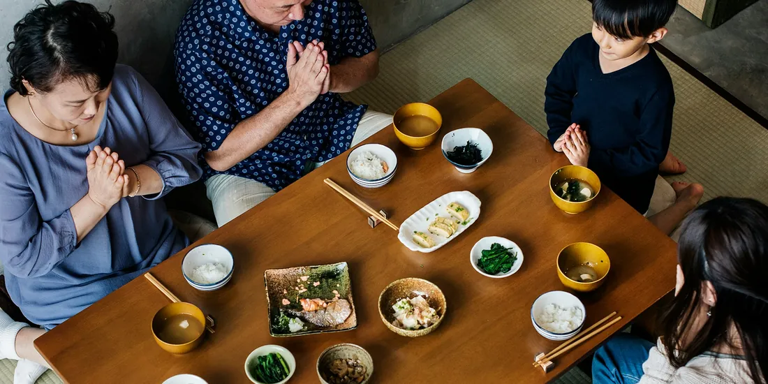 畳に置かれた低い木の卓を三世代の家族が囲み、食事の前に手を合わせている様子を真上から。