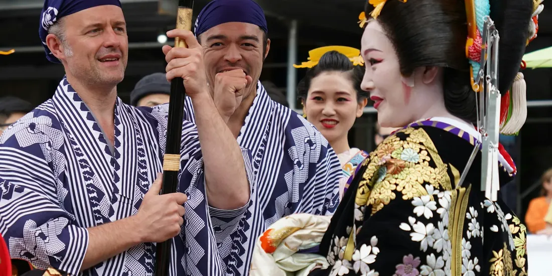 東京の夏祭りで、藍の浴衣姿の二人が伝統衣装の演者と笑い合っている様子。