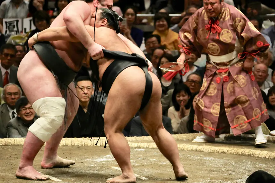 Two sumo wrestlers locked together in the ring as the referee calls the bout.