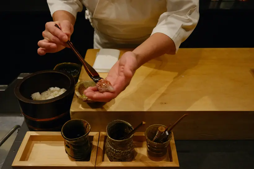 A chef's hands shaping nigiri and brushing it with sauce at a counter.