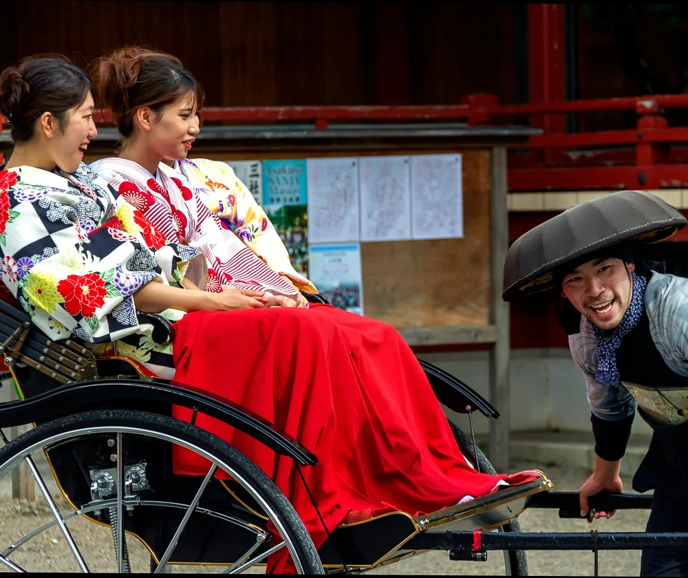 Two guests in kimono riding a jinrikisha past Senso-ji while the driver talks with them.