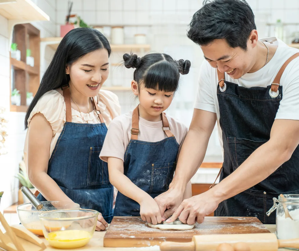 A family making food together at a kitchen counter, the child shaping dough between her parents.